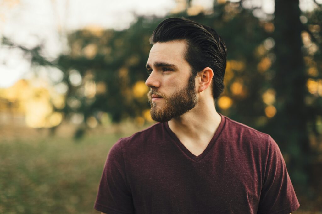 A bearded young man in a burgundy shirt standing in nature and looking to the side