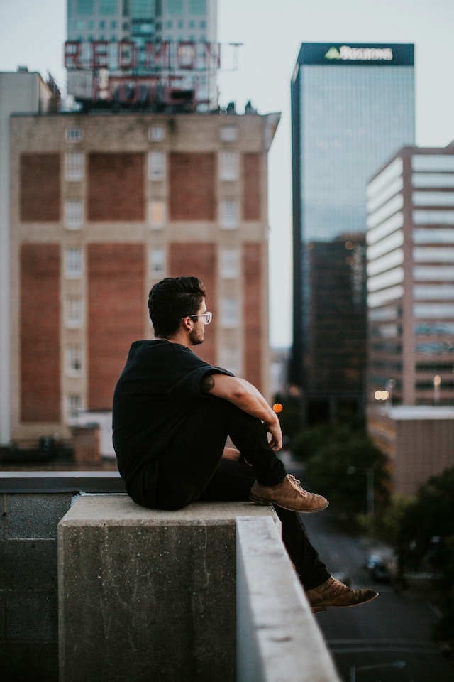 a man sitting high up on a concrete balcony edge with tall cityscape buildings in the background