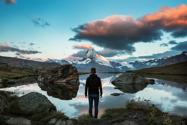 A man standing on the bank of a river looking towards snowy mountains in the distance