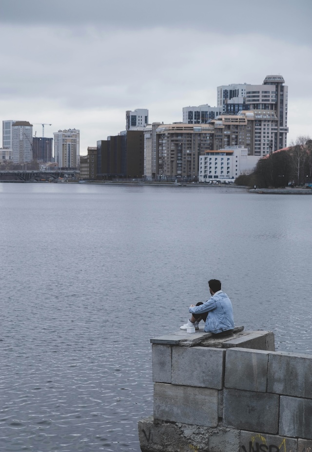 A man wearing a jean jacket sitting on a concrete structure overlooking water with city buildings on the other side of the bay